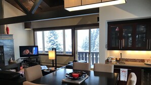 A modern living room with a large flat-screen TV, a dining table with chairs, a fireplace, and a kitchen area with cabinets and a sink.