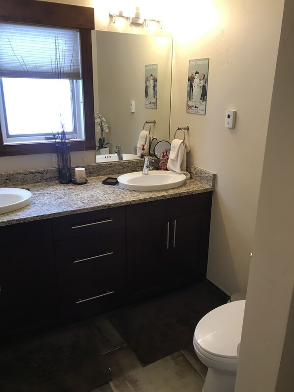 A bathroom with a double sink, dark cabinets, a toilet, a window with blinds, and a mirror with a poster. - Beautiful Vail Home-Close to Free Vail Bus, Skiing and Golf! (Vail)