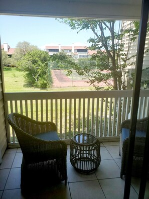 A balcony with wicker furniture, a glass table, and a view of a tennis court and buildings.