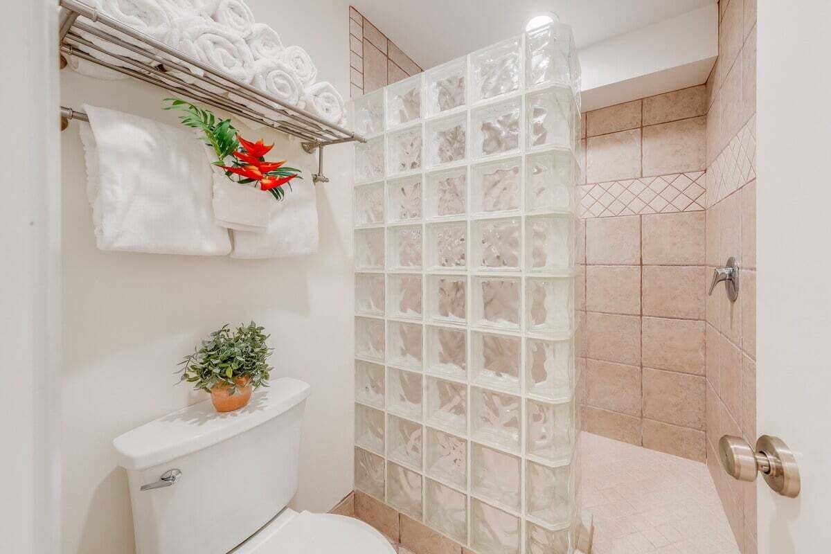 Ein Badezimmer mit Duschkabine aus Glas, Toilette, Waschbecken mit Topfpflanze und Handtuchhalter mit aufgerollten Handtüchern.
