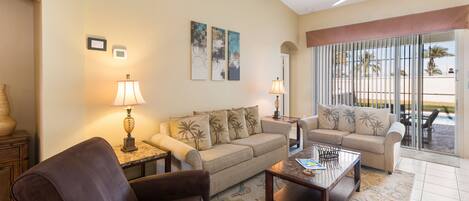 A living room with a beige sofa, two armchairs, a coffee table, and a ceiling fan. There are two wall-mounted lamps and a large window with a view of palm trees.