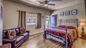 A bedroom with a bed, a wooden floor, a ceiling fan, a window with curtains, a blue door, and a brown leather sofa. - Historic San Jose House Room 2 Wyatt Earp (Tombstone)