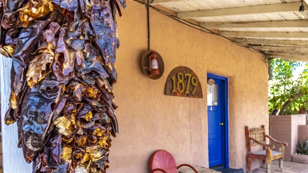 A rustic building exterior with a blue door, a vintage chair, and a hanging dried plant. - Stay at the Historic San Jose House Room 4 Doc Holliday (Tombstone)