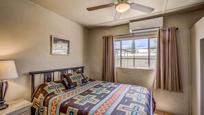 A bedroom with a bed, a ceiling fan, an air conditioning unit, a window with curtains, and a nightstand with a lamp. - Stay at the Historic San Jose House Room 4 Doc Holliday (Tombstone)