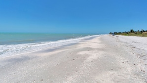 A wide sandy beach with gentle waves, clear skies, and a few people in the distance.