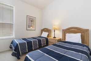 Quarto com duas camas de solteiro, mesa de cabeceira com abajur, quadro emoldurado na parede e janela com persianas.