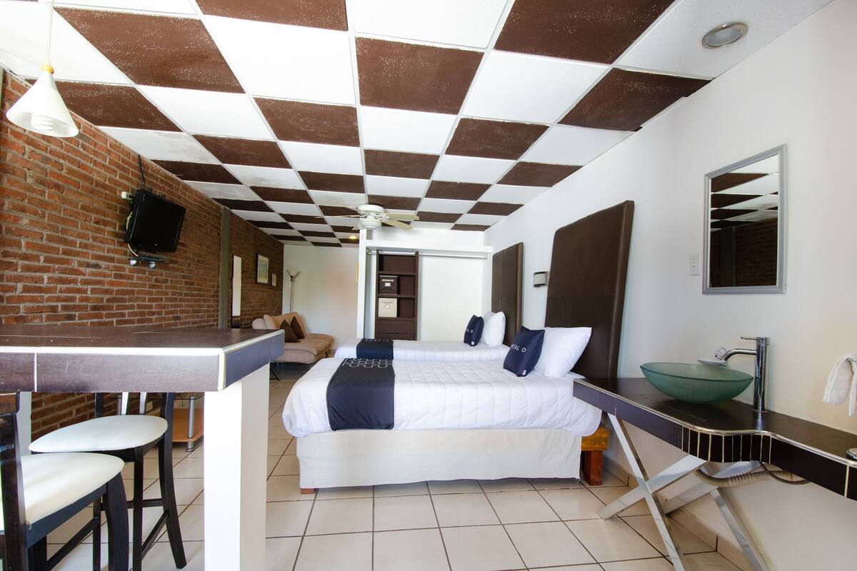 A hotel room with a checkered ceiling, a bed with white bedding and dark pillows, a green bowl sink, and a bar area with stools.