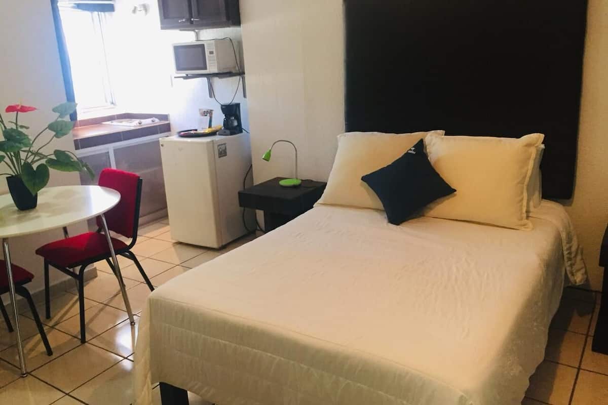 A hotel room with a bed, a small desk, a chair, a table, and a potted plant.