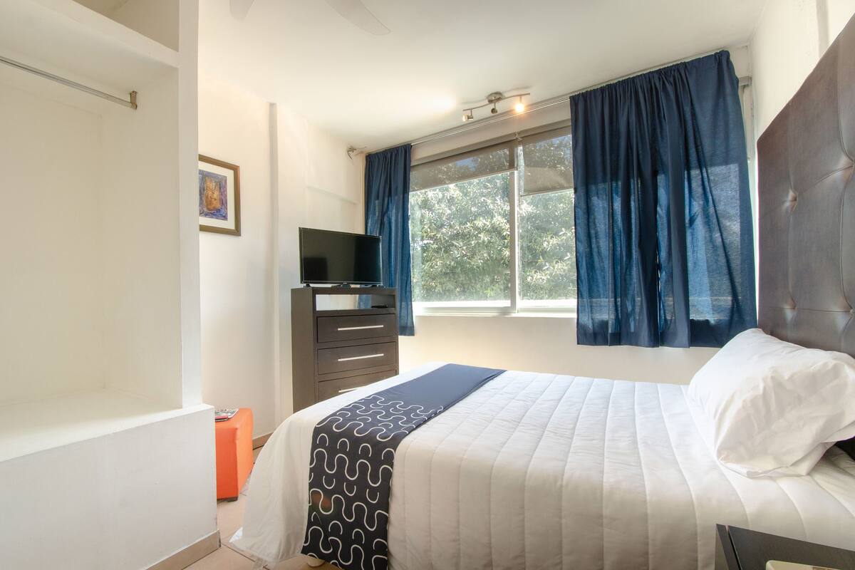A bedroom with a bed, a TV, a dresser, and a window with curtains.