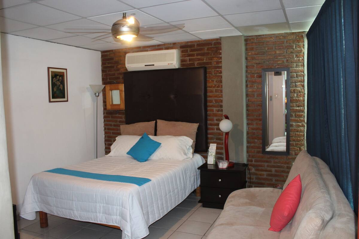 A bedroom with a bed, a nightstand, a sofa, a mirror, and a ceiling fan.