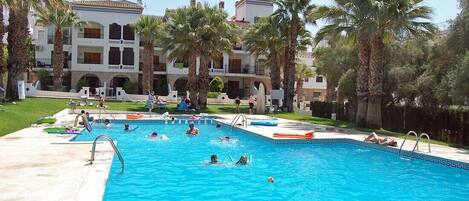 A swimming pool with multiple lanes, surrounded by palm trees and a large building with balconies.