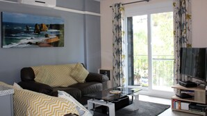 A living room with a sofa, coffee table, and TV. There is a painting on the wall and a window with curtains.