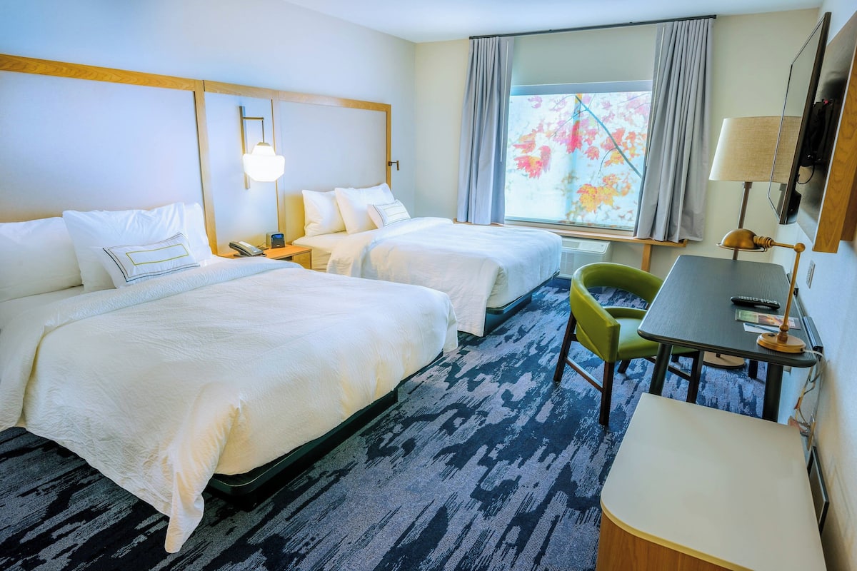 A hotel room with two beds, a desk, a chair, a lamp, and a window with curtains.