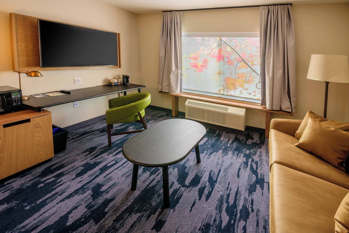 A hotel room with a desk, a green chair, a sofa, a TV, and a window with curtains.