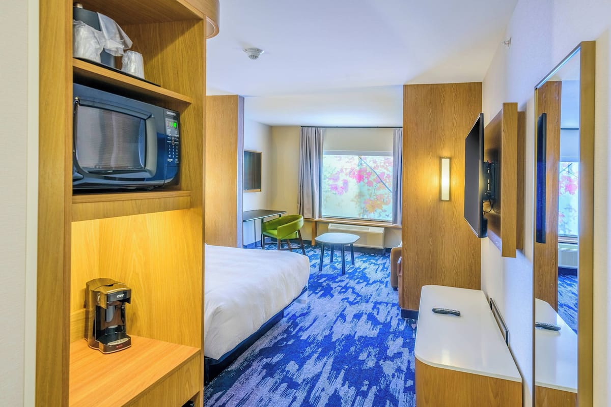 A hotel room with a bed, a microwave, a coffee maker, a desk, a chair, a small table, and a TV.