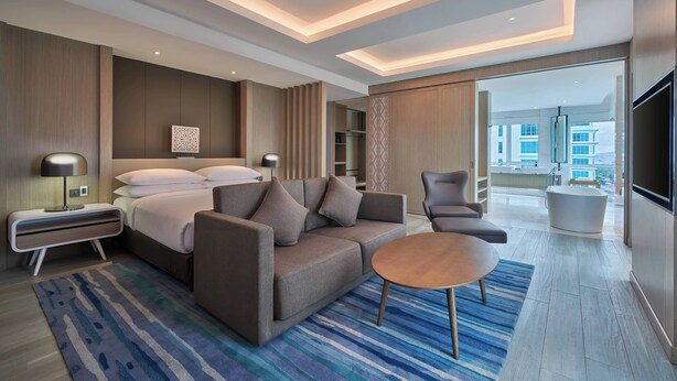 A modern hotel room with a bed, sofa, armchair, and a small table. There is a large window with a view of buildings. - Fairfield by Marriott Bintulu Paragon (Bintulu)