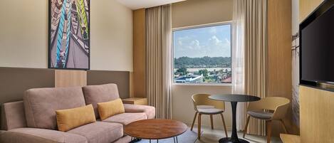 Quarto de hotel moderno com sofá, mesa redonda pequena, duas cadeiras, janela grande com vista e televisão de tela plana.