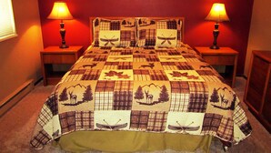 A bed with a quilt featuring animal and boat patterns, flanked by two bedside lamps, in a room with red walls. - Paintbrush North- clean & quiet home with mountain views (West Yellowstone)