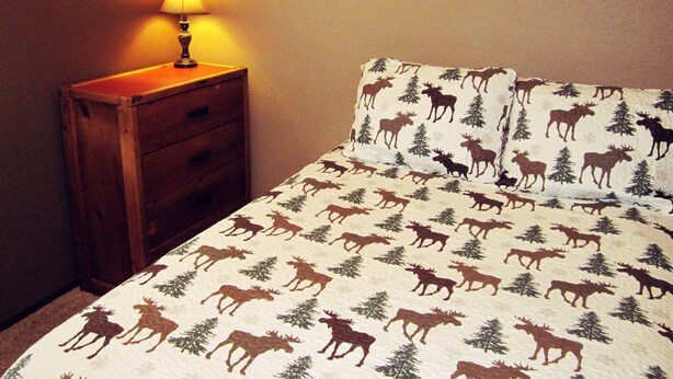 A bedroom with a bed featuring a moose and pine tree pattern, a wooden nightstand with a lamp, and a wall with a warm light. - Paintbrush Grande - spacious & comfortable, perfect for families (West Yellowstone)