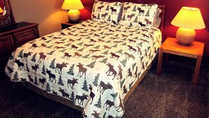 A bedroom with a bed featuring a moose and tree pattern, two bedside lamps, a framed picture on the wall, and a wooden nightstand. - Sandhill East Home - Mountain Views, Portable A/C & 15 Minutes to Yellowstone (West Yellowstone)