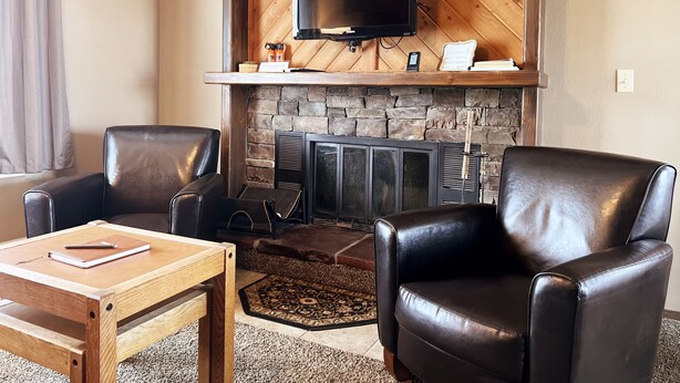 A cozy living room with a fireplace, leather chairs, a wooden coffee table, and a flat-screen TV mounted on a wooden wall. - Sandhill East Home - Mountain Views, Portable A/C & 15 Minutes to Yellowstone (West Yellowstone)