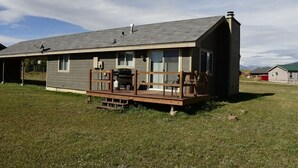 A single-story house with a deck, a chimney, and a satellite dish. - Sandhill East Home - Mountain Views, Portable A/C & 15 Minutes to Yellowstone (West Yellowstone)