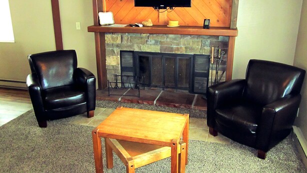 A living room with two leather chairs, a wooden coffee table, a fireplace with a flat-screen TV above it, and a clock on the wall. - Sandhill West Home - comfortable family retreat with great views (West Yellowstone)