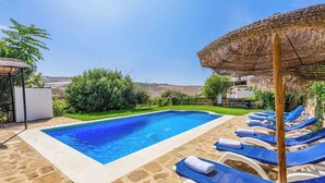 Ein rechteckiger Swimmingpool mit klarem, blauem Wasser, umgeben von Steinplatten und einer weißen Umrandung. Blaue Liegestühle mit weißen Handtüchern, ein strohgedeckter Sonnenschirm und im Hintergrund eine Hügellandschaft.
