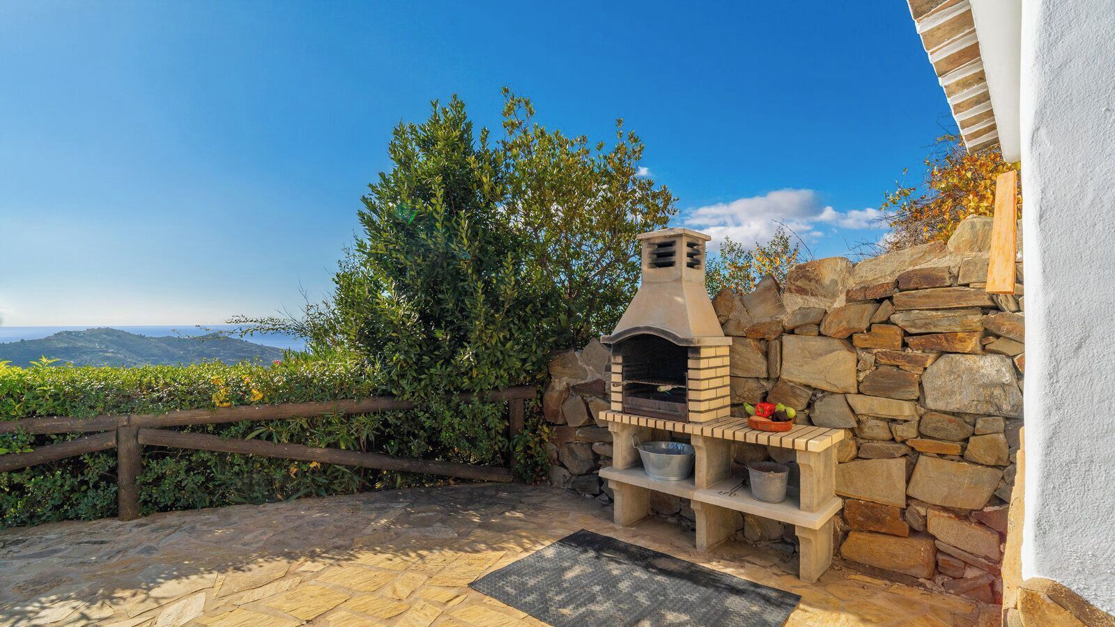 A stone-built outdoor oven with a stone wall backdrop, a wooden fence, and a view of distant hills.