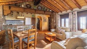 A rustic interior with a wooden dining table, chairs, a sofa, and a kitchen area with a refrigerator. - Finca Algarabia Cómpeta by Ruralidays (Málaga)