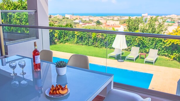 Ein Balkon mit einem Glastisch, zwei Weingläsern, einer Flasche Rotwein, einer Obstplatte und Blick auf einen Swimmingpool und Grünflächen.