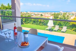 Ein Balkon mit einem Glastisch, zwei Weingläsern, einer Flasche Rotwein, einer Obstplatte und Blick auf einen Swimmingpool und Grünflächen.