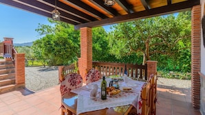 A covered patio with a table set for dining, surrounded by brick pillars and wooden railings, overlooking a lush garden and distant hills. - La Recacha Órgiva by Ruralidays (Granada)