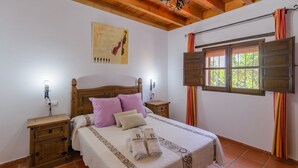 A bedroom with a wooden bed, two bedside tables, a nightstand, a window with curtains, and a wall-mounted artwork. - La Recacha Órgiva by Ruralidays (Granada)
