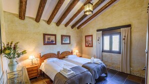 A room with two beds, wooden beams on the ceiling, a window with curtains, and a vase with flowers on a table.
