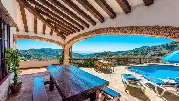 A terrace with a pool, wooden furniture, and a view of the hills.
