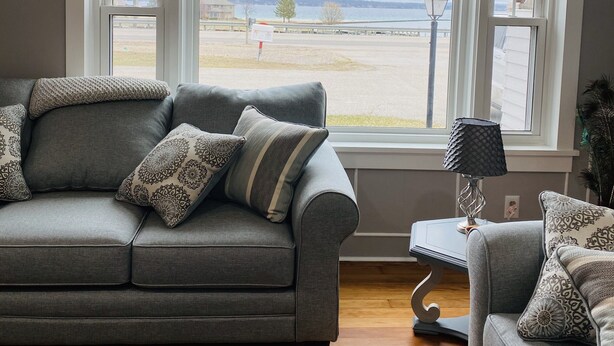 Una sala de estar con un sofá gris, cojines estampados, una mesita con una lámpara y vistas a una playa y al agua a través de ventanas amplias.