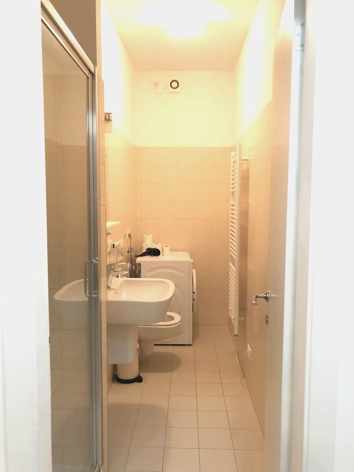 A small, narrow bathroom with a glass shower enclosure, a sink, and a toilet.