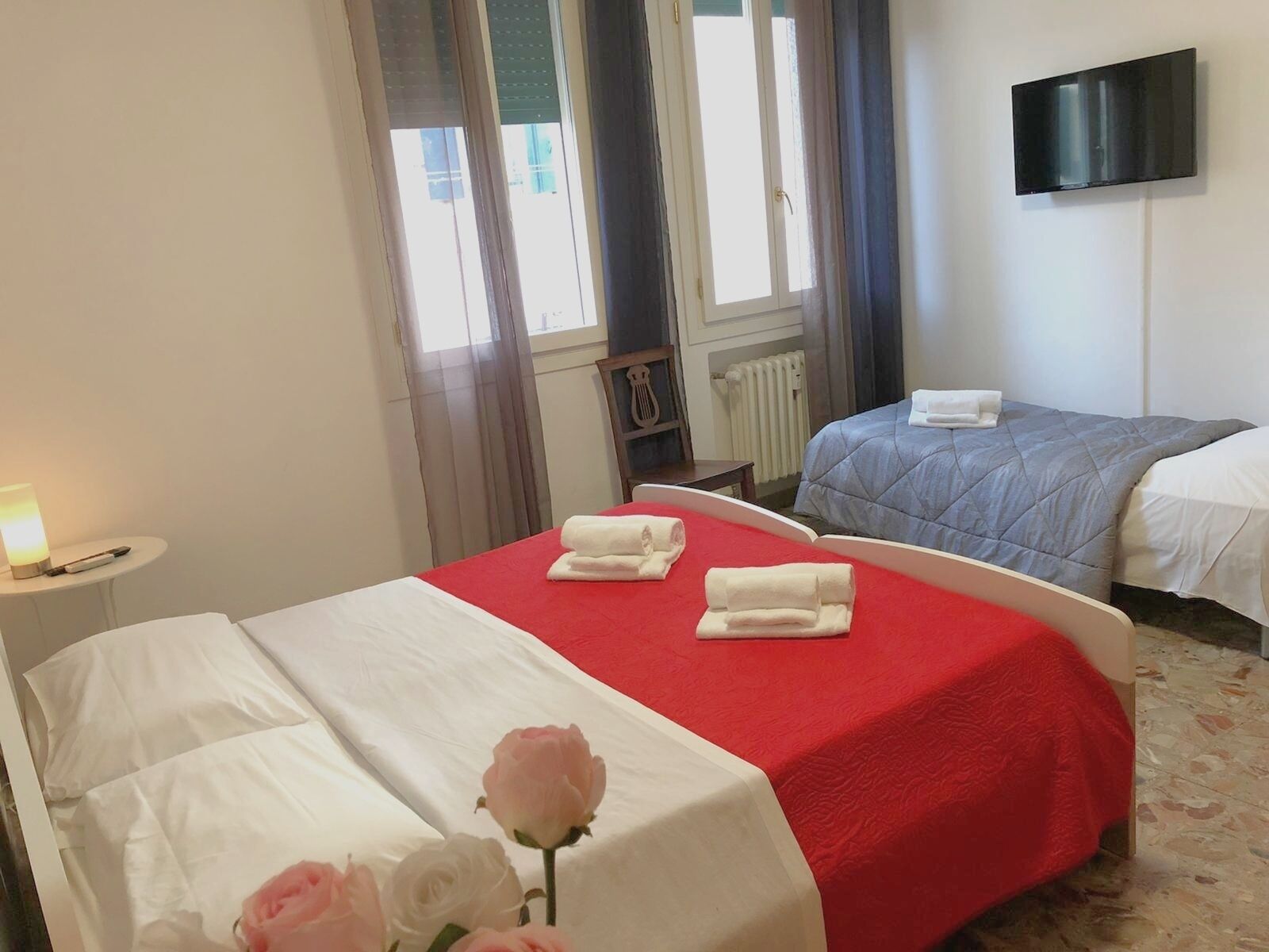 A hotel room with two beds, one with a red bedspread and the other with a blue bedspread. There is a TV mounted on the wall, a small table with a lamp, and a radiator.
