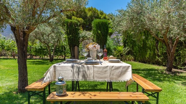 Una mesa de comedor al aire libre en un jardín, con mantel, platos, vasos y una botella.