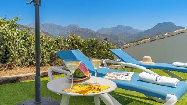 A sunlit outdoor seating area with a mountain backdrop, a table with a glass of wine and a watermelon slice, and a towel with a logo.