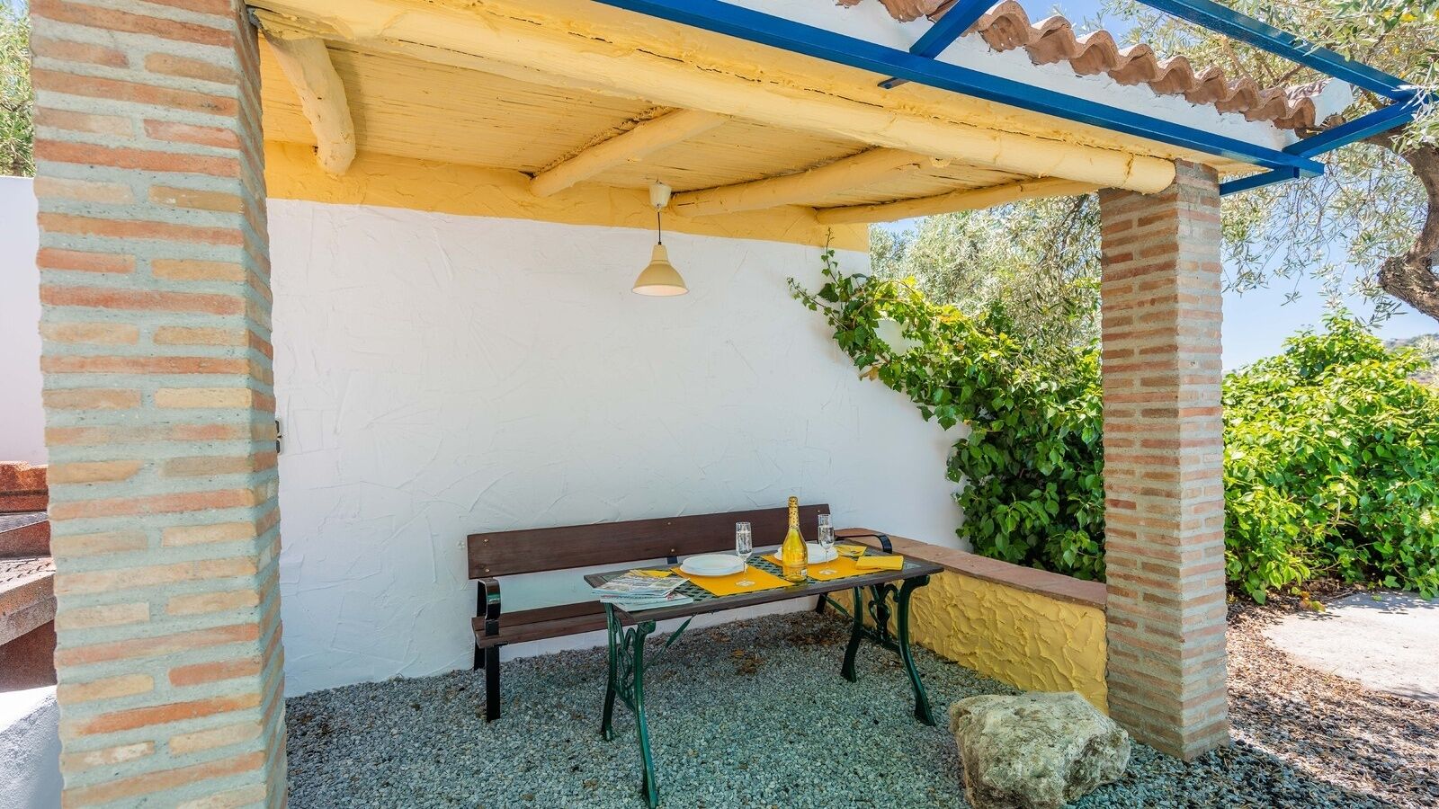 Un área de estar al aire libre con un banco y mesa, rodeada por un muro amarillo y follaje.