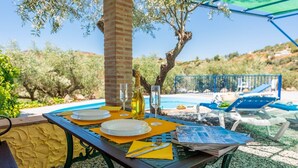 Une table à manger installée à l’extérieur, avec des assiettes, des couverts et une bouteille de vin, et en arrière-plan, une piscine et des chaises longues.