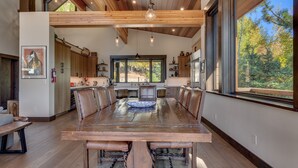 A spacious dining area with a large wooden table and leather chairs, a kitchen with wooden cabinets, and large windows offering a view of the outdoors.