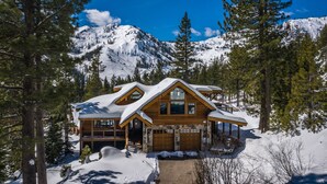 A wooden cabin with a snow-covered roof, surrounded by pine trees and a mountain backdrop.