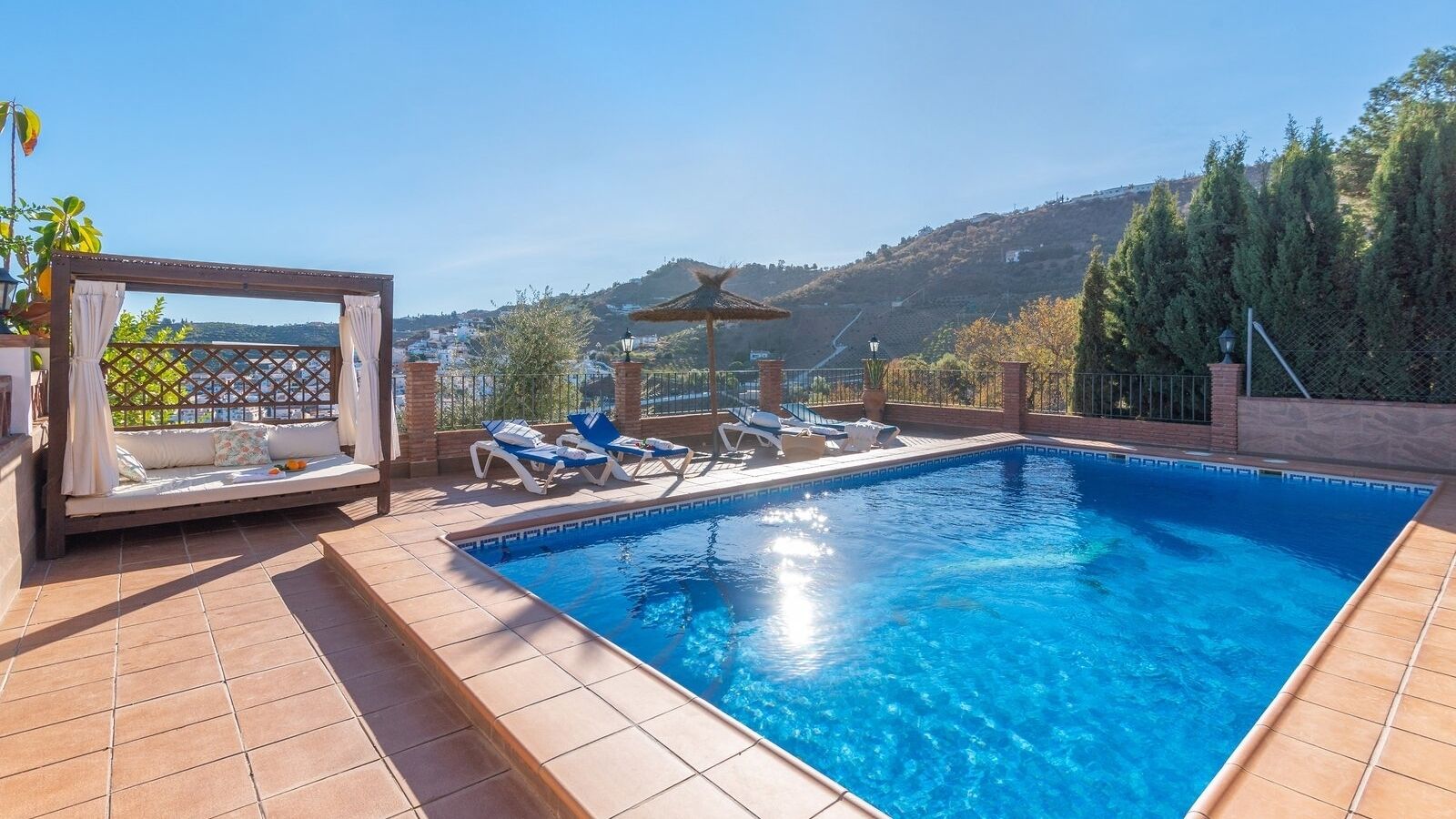 A rectangular swimming pool with clear blue water, surrounded by tiled flooring. There are lounge chairs and a covered seating area with a view of the hills in the background.