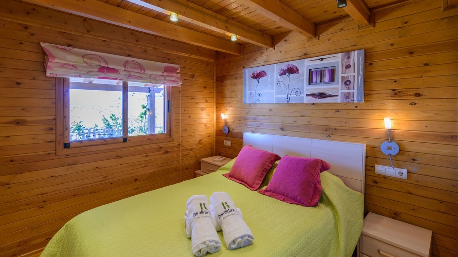 A wooden cabin room with a bed, two pillows, a window, and a wall-mounted light fixture.