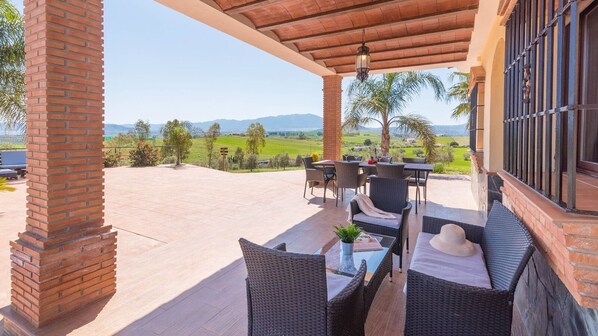 Un porche cubierto con mobiliario exterior, un pilar de ladrillo y vistas a un paisaje verde.