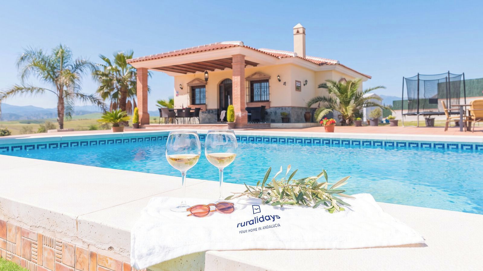 Una piscina amb dos gots de vi blanc i una tovallola, una casa amb un patí, i un rètol que diu 'ruralidays your home in Andalucia'.