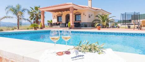 A pool with two glasses of white wine and a towel, a house with a patio, and a sign that reads 'ruralidays your home in Andalucia'.